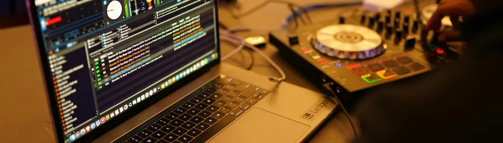 Laptop with audio editing software interface, audio mixer, and recording equipment on a cluttered desk.