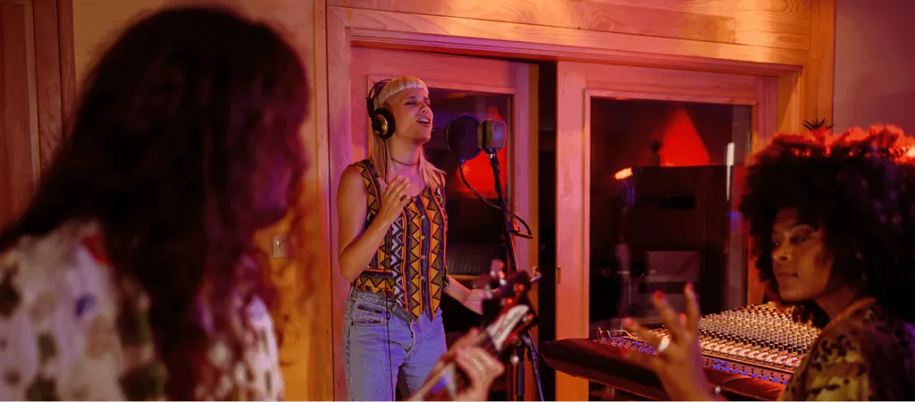Woman with headphones singing in recording studio with orange lighting, another person visible at mixing board in background.