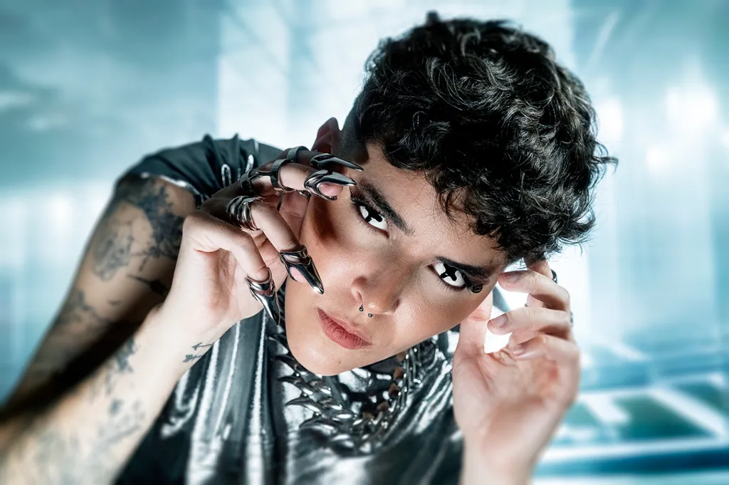 Person with dark curly hair and tattoos holding black sunglasses, looking at camera against blue-tinted background.