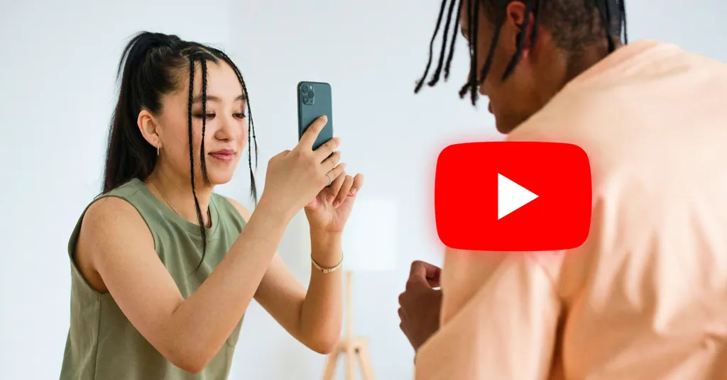 Woman in olive tank top holds phone filming shirtless man with dreadlocks. Red YouTube play button logo overlaid in center.