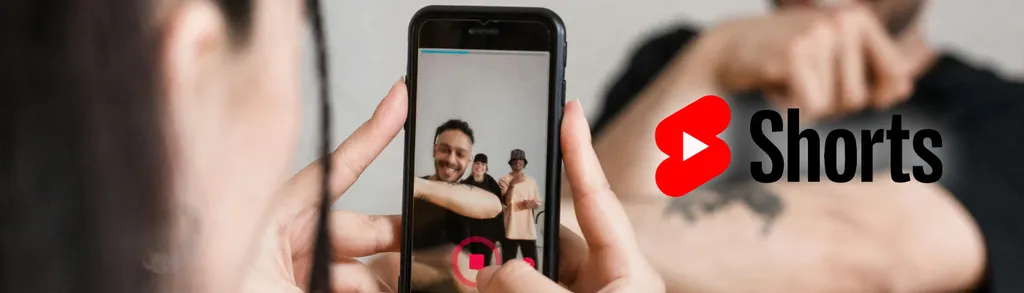 Mani tengono smartphone che riprende video, schermo mostra due persone, logo YouTube Shorts rosso e nero a destra.