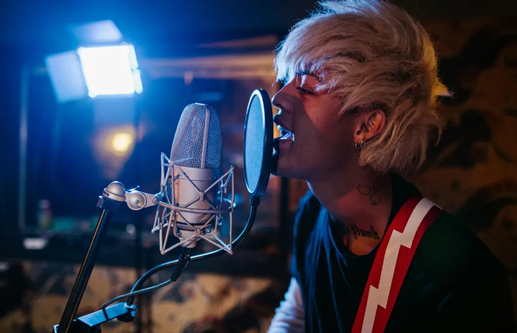 Hombre mayor con gafas cantando en micrófono de estudio, iluminación azul, cabello rubio, camiseta con rayas rojas y blancas.