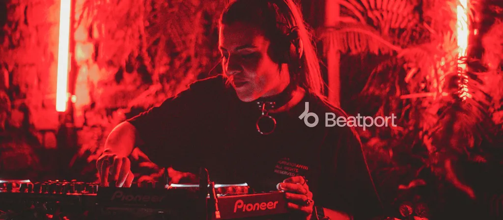 DJ performing at mixing console in dark venue with dramatic red lighting and decorative backdrop.