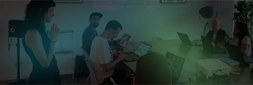 Dimly lit office scene, multiple people working at desks with laptops, desk lamps and plants visible.