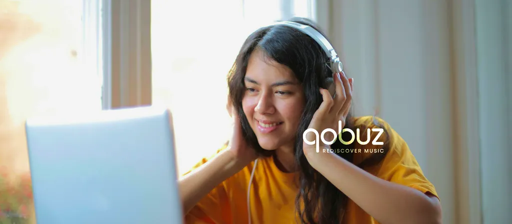 Woman in yellow shirt wearing headphones while using laptop computer indoors with natural lighting from window.